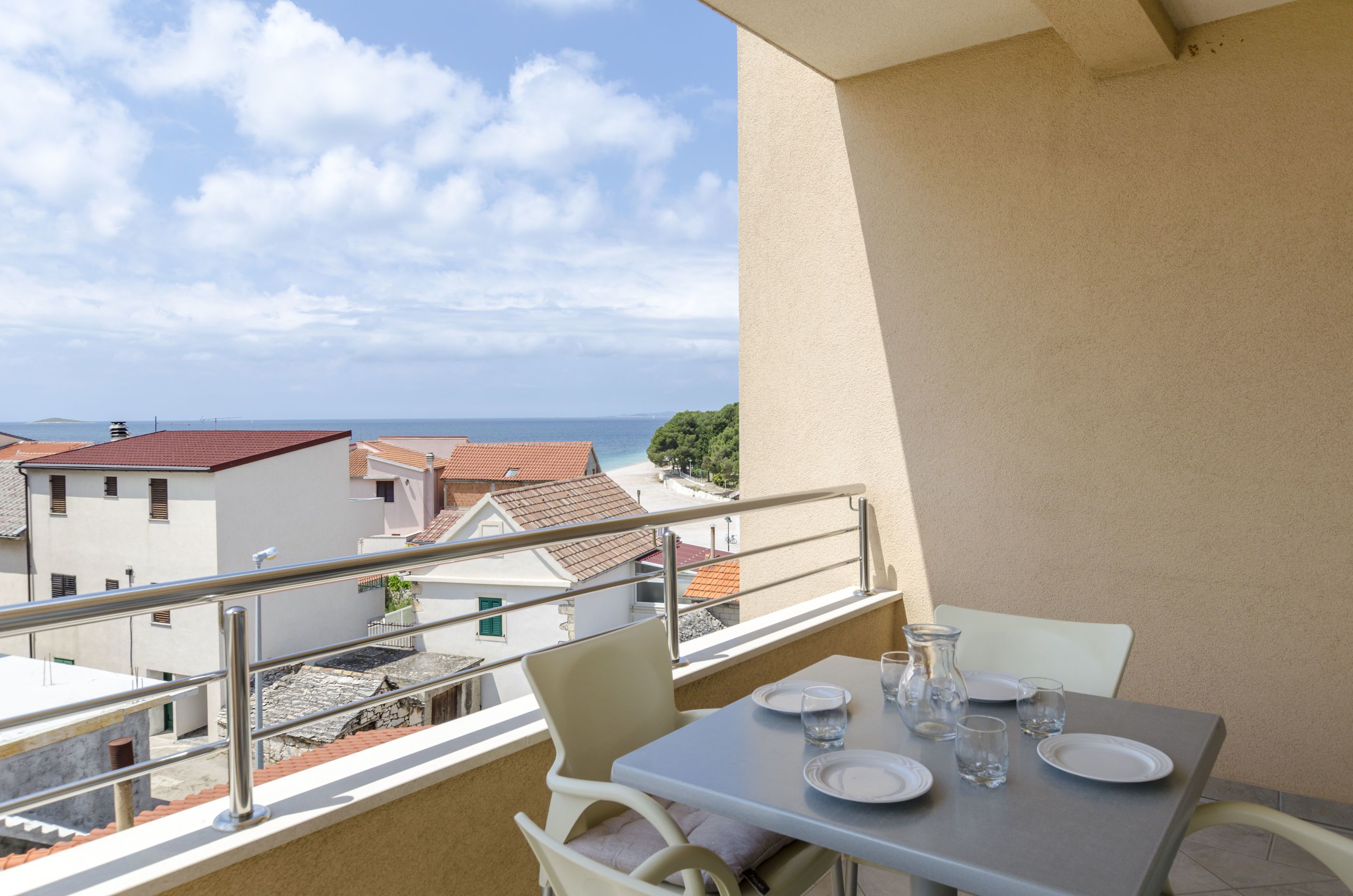Un balcón con una mesa lista para dos personas, con vistas a la costa, edificios y al mar.
