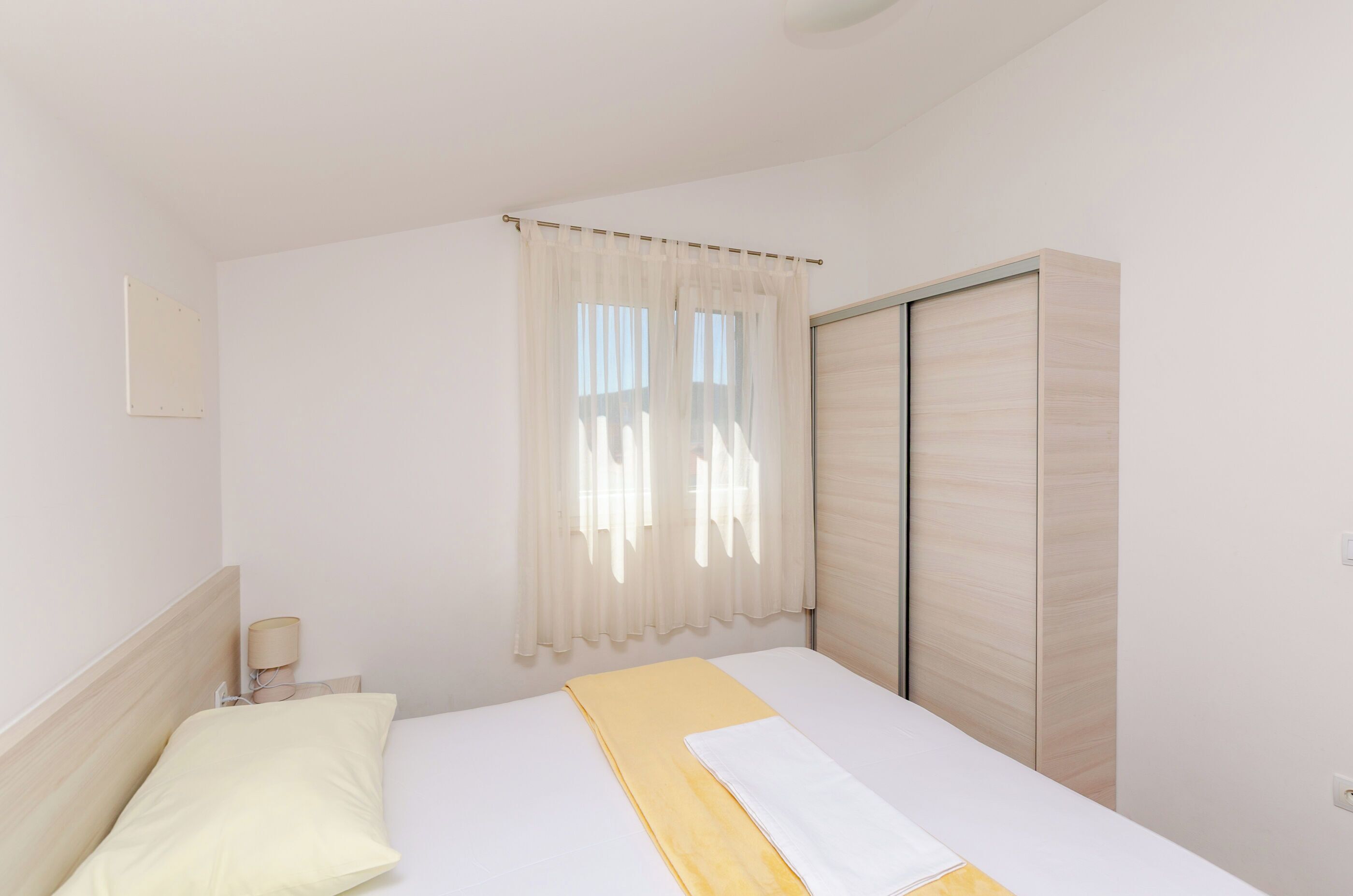 A bedroom with a bed, a nightstand, a lamp, a window with sheer curtains, and a wardrobe.