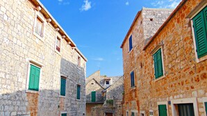A narrow cobblestone street flanked by historic stone buildings with green shutters, leading to a central building with a pitched roof. - Apartments Mare (Postira)