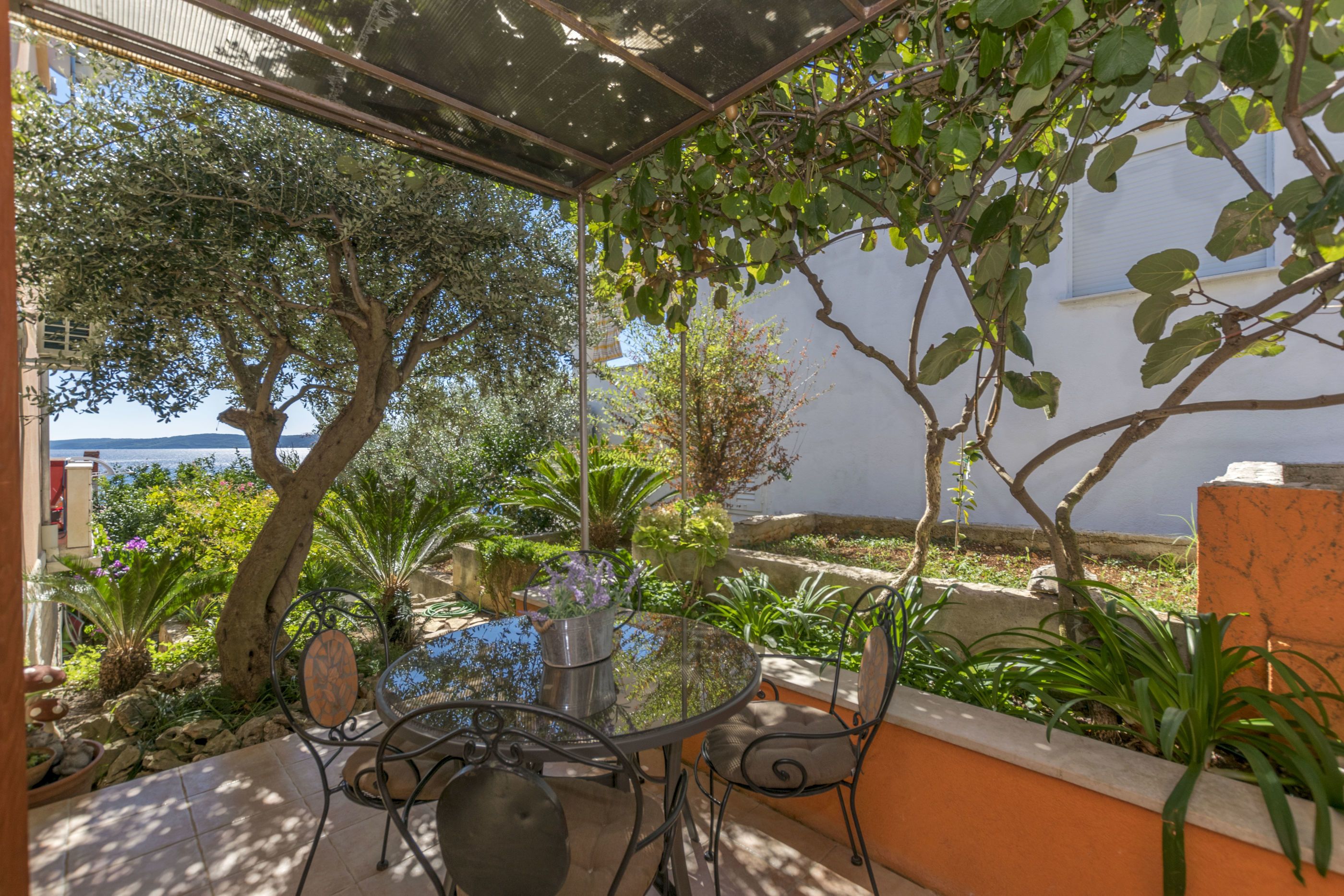 A covered outdoor patio with a table and chairs, surrounded by various plants and trees, overlooking a body of water.