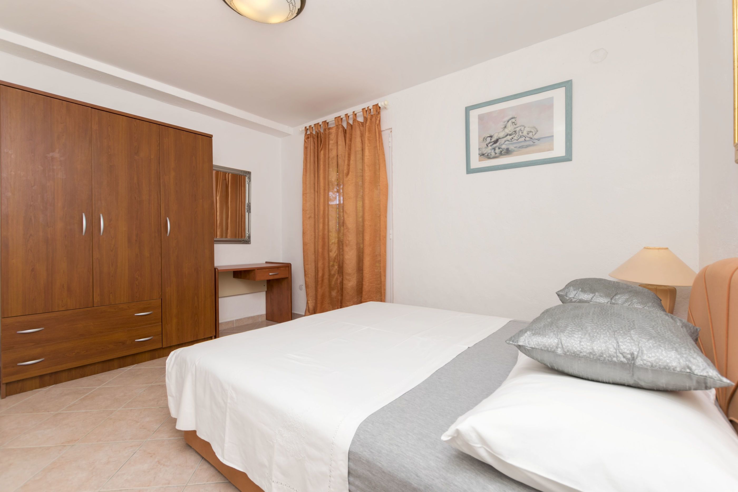 Een slaapkamer met een bed, een houten garderobe, een nachtkastje, een lamp en een ingelijst schilderij aan de muur.