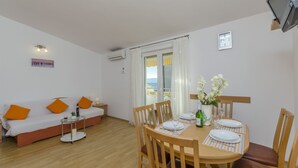 A modern living room with a sofa, dining table, and chairs. There is a window with curtains, a clock on the wall, and an air conditioning unit. - Apartments Srecko (Duce)