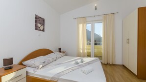A neatly made bed with a floral bedspread, a bedside table with a lamp, and a view of a balcony with curtains. - Apartments Srecko (Duce)