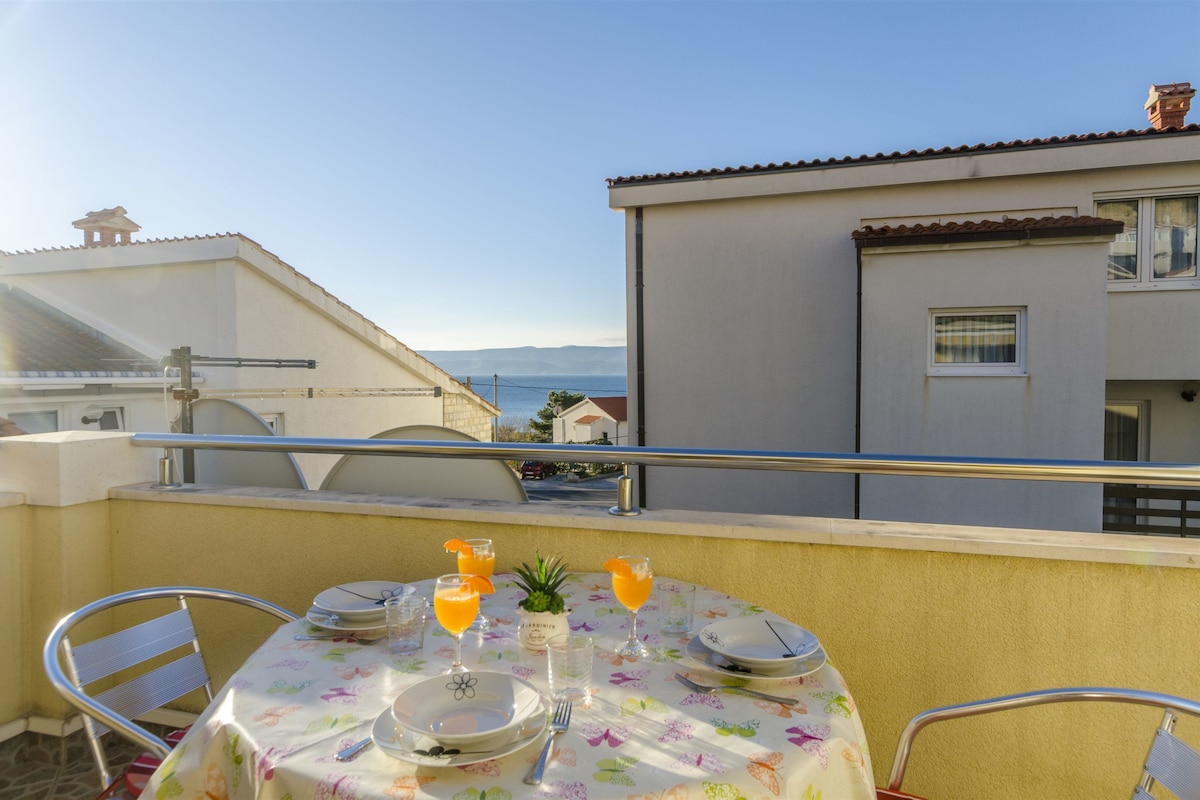 A balcony with a table set for two, overlooking a residential area and the sea.