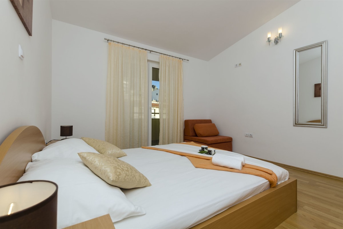 A hotel room with a large bed, a chair, a mirror, and a view of the outside.