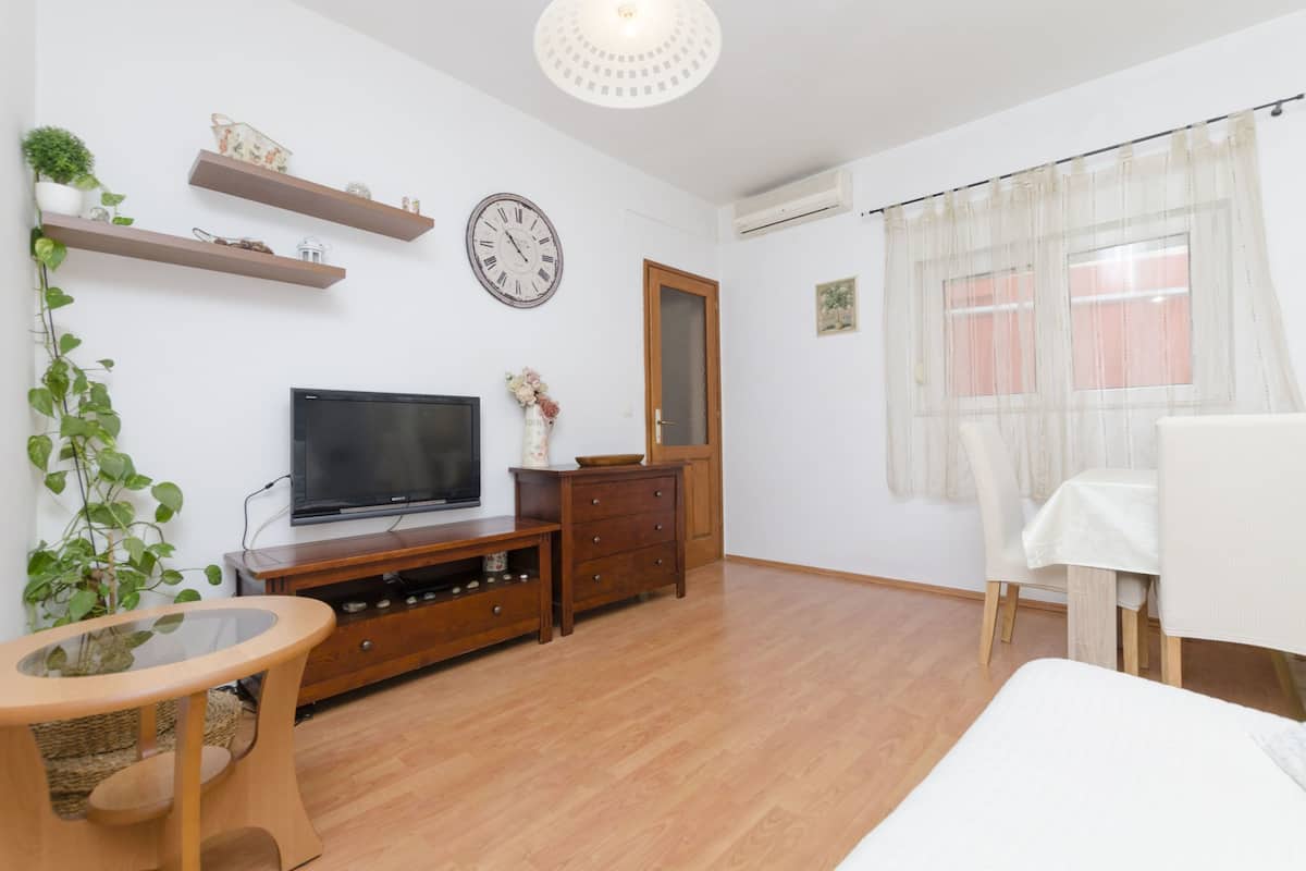 A living room with a TV, wooden furniture, a clock, and a window with sheer curtains.