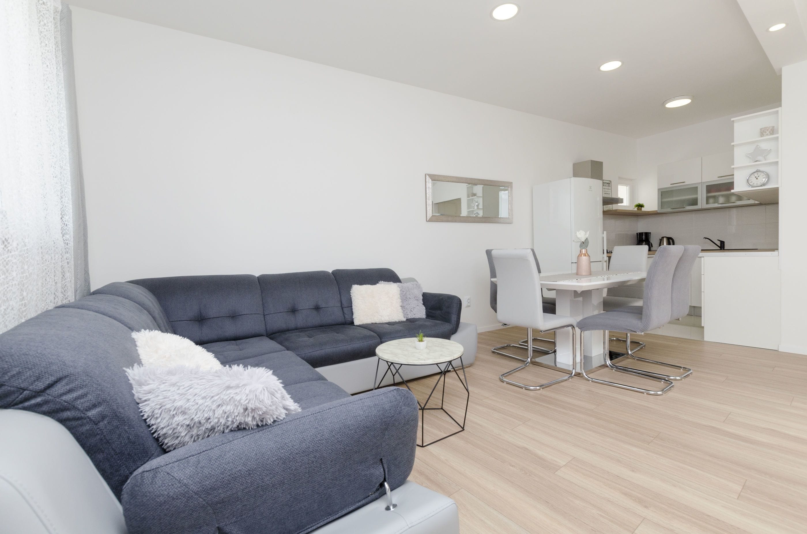 Una sala de estar moderna con un sofá seccional gris, una mesa de centro redonda y una zona de comedor con una mesa y sillas blancas.