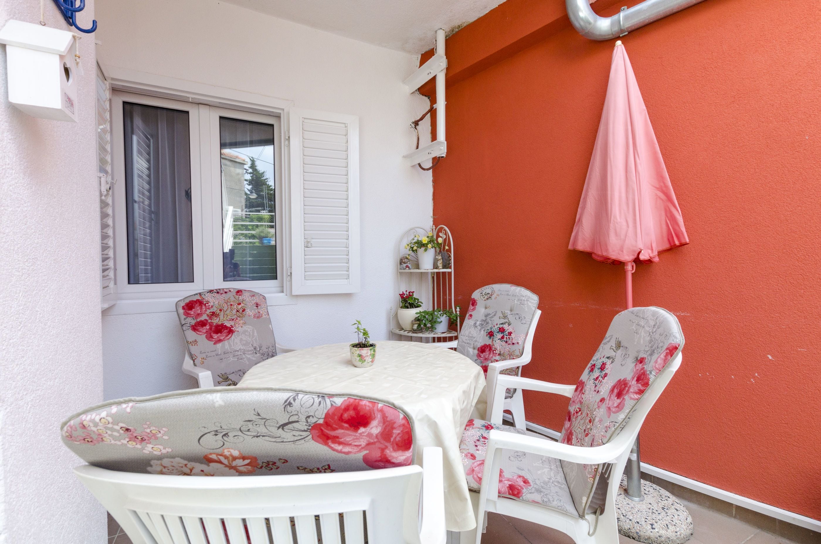 A small patio area with a table and chairs, a window with shutters, a potted plant, and a pink umbrella.