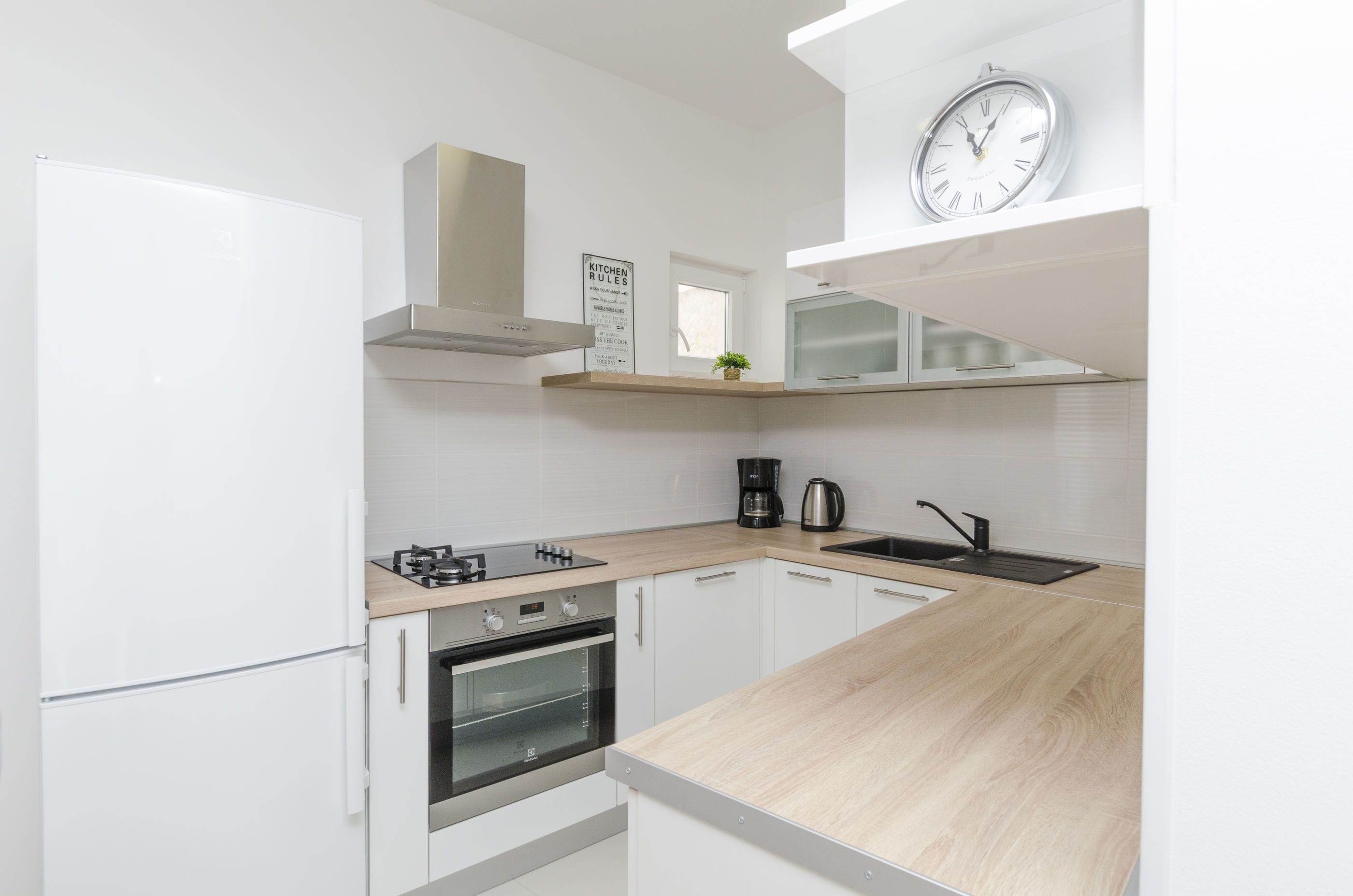 A modern kitchen with white cabinetry, a wooden countertop, a built-in oven, a coffee maker, and a clock on the wall.
