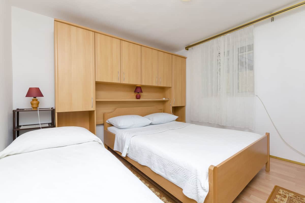 A bedroom with two single beds, wooden wardrobe, bedside tables, and a window with sheer curtains.