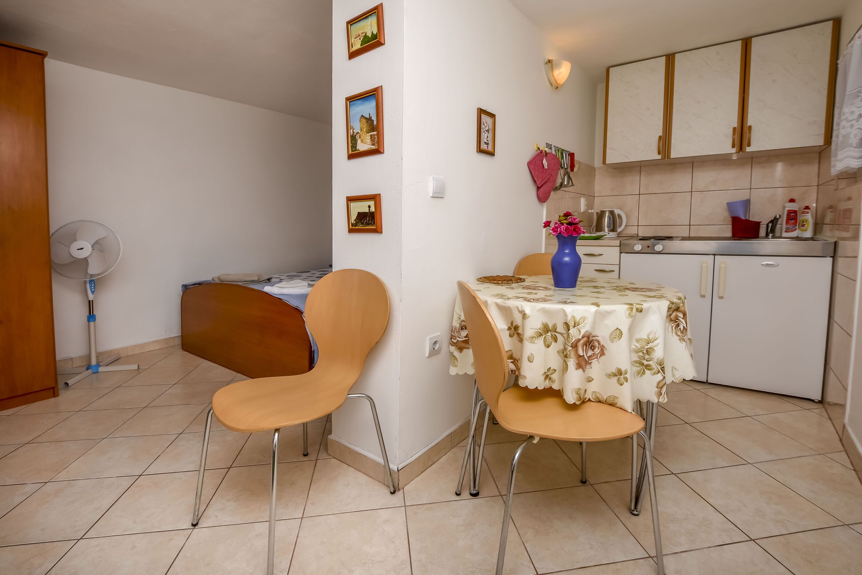 A dining area with two wooden chairs, a table with a floral tablecloth, a fan, and a kitchen with cabinets and a stove.