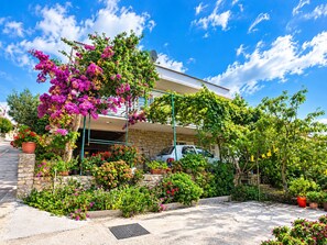 A residential building with a stone facade, a car parked in the driveway, and a variety of flowering plants and shrubs. - Apartment Anka (Primosten)