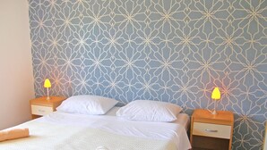 Una cama bien hecha con sábanas blancas y una toalla enrollada, dos mesitas de noche con lámparas, y una pared con estampado.