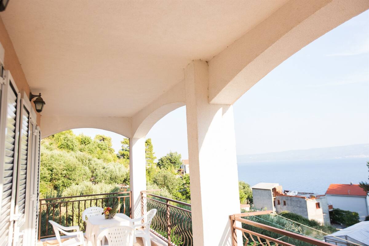 A balcony with white chairs and a table, overlooking a scenic landscape with trees and a view of the sea.