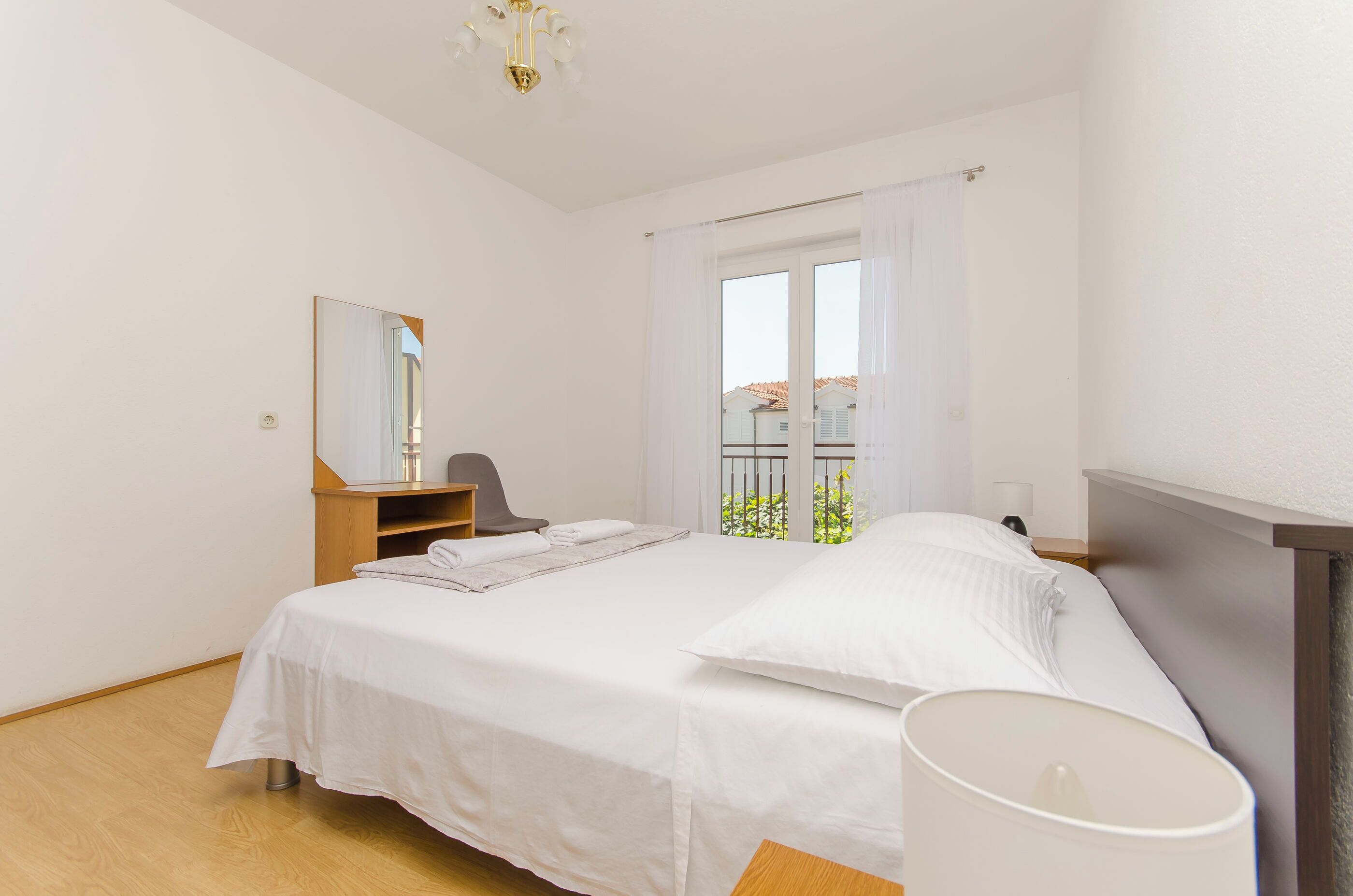 Quarto com cama, cadeira, escrivaninha, espelho e janela com vista para a varanda.