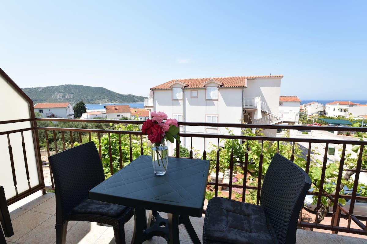 A balcony with a table and chairs, a vase with flowers, and a view of buildings and the sea.