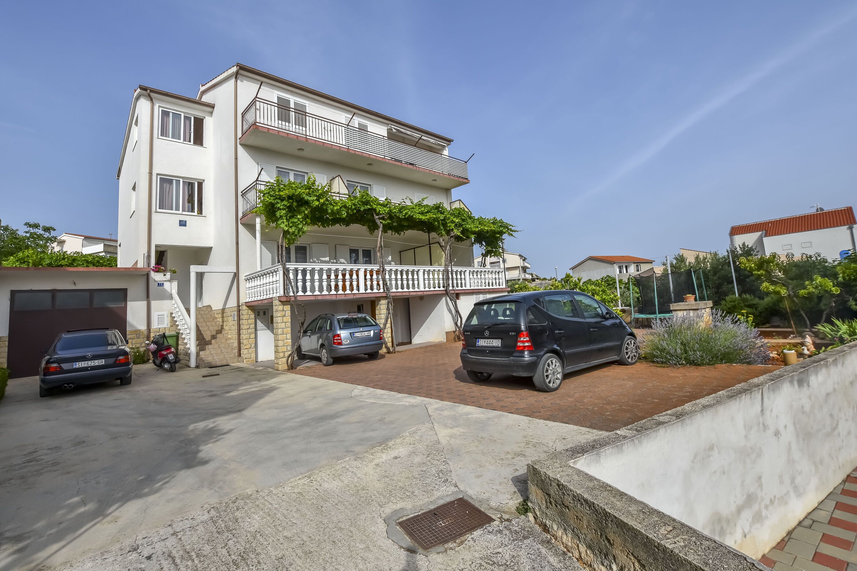 Un edificio residenziale a due piani con un balcone, auto parcheggiate e un’area giardino.