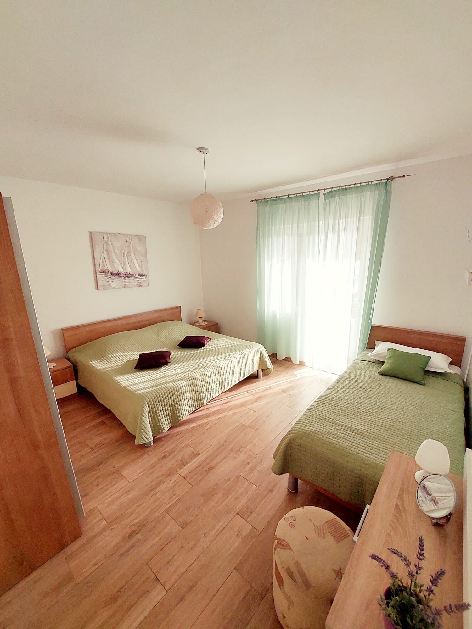 Ein Zimmer mit zwei Betten, Holzboden, einem Bild an der Wand und einem Fenster mit Vorhängen.