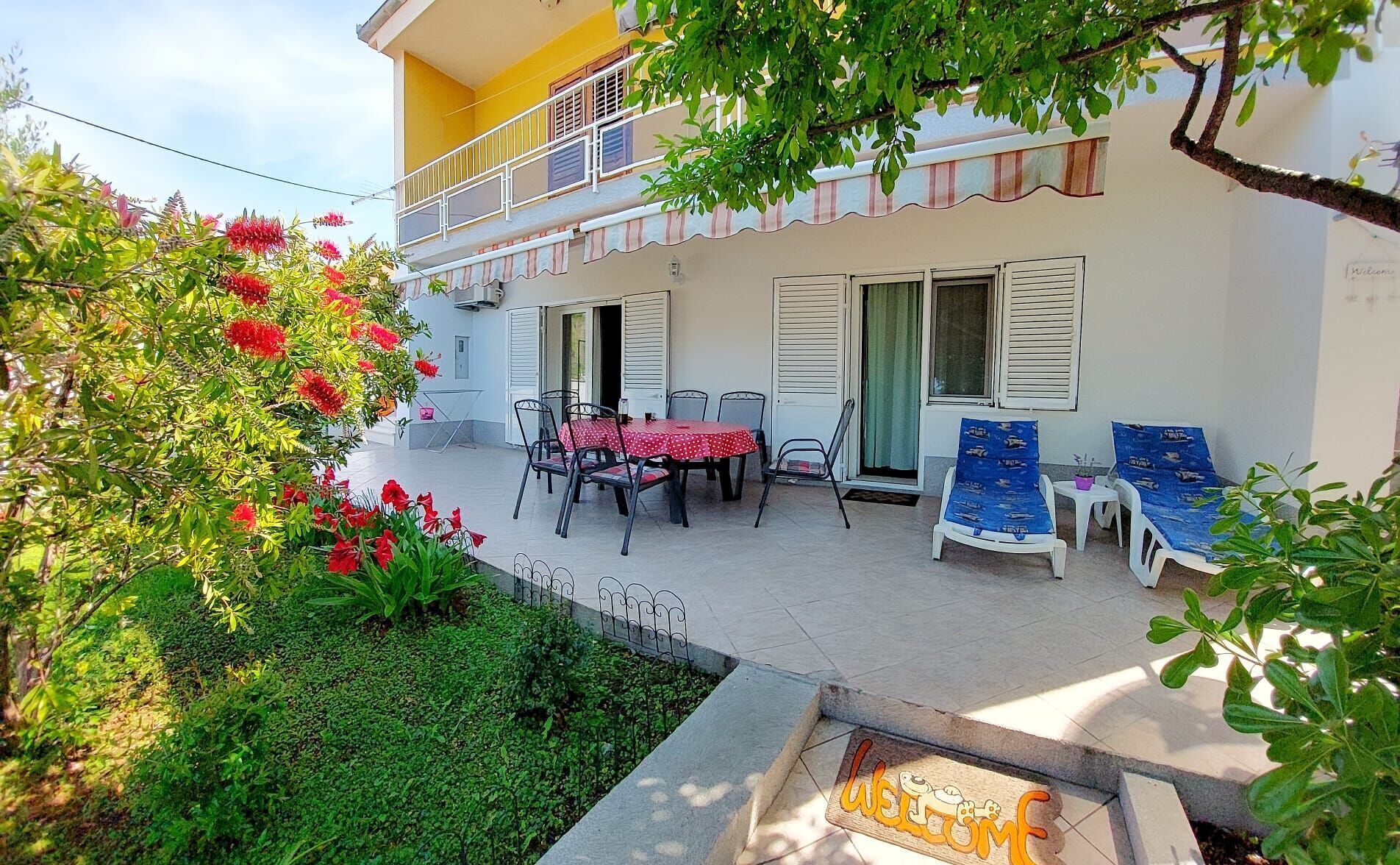 A two-story building with a balcony, a patio with a table and chairs, and a welcome mat with a dog illustration.