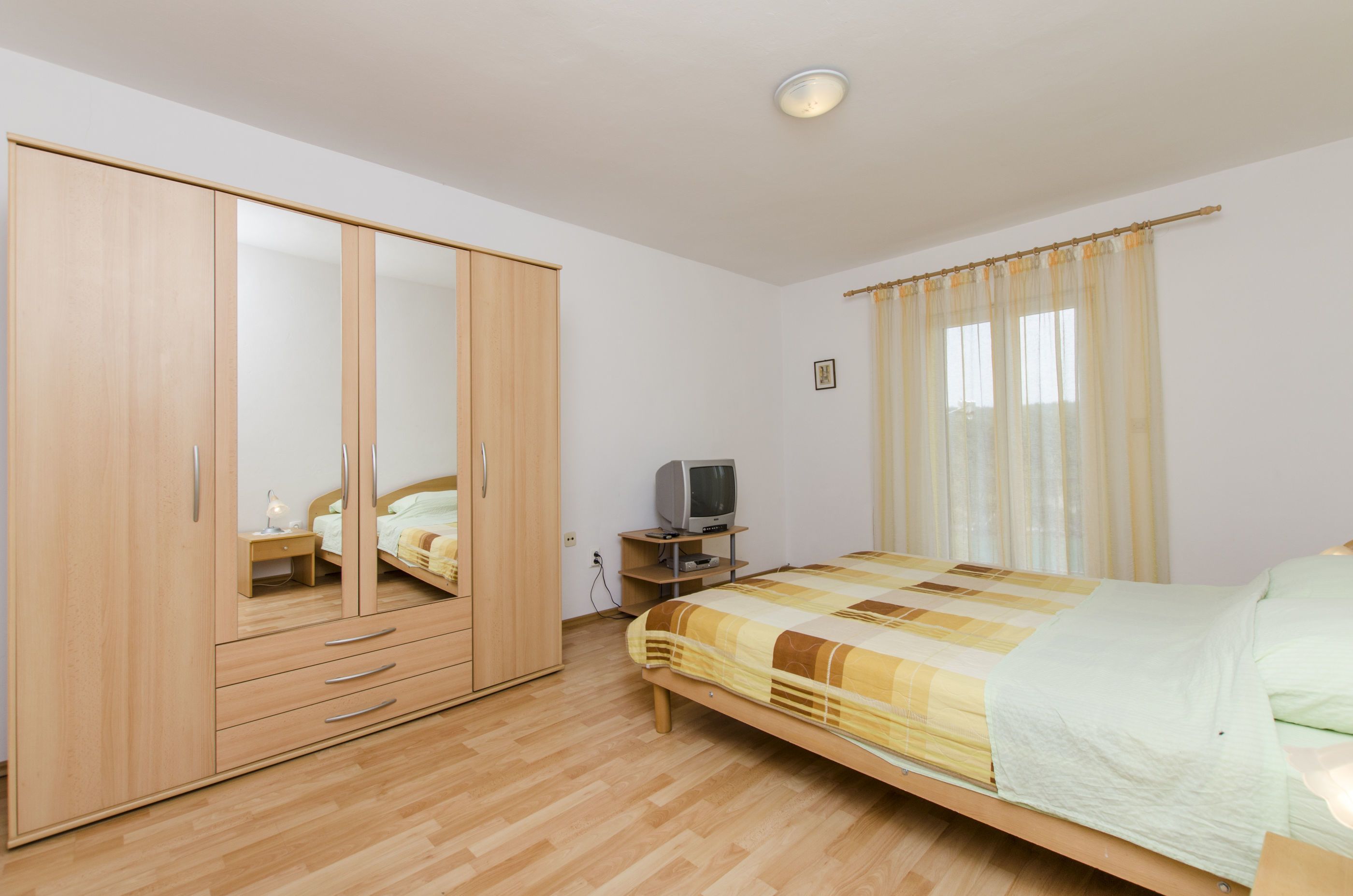 A bedroom with a wooden wardrobe, a bed with a checkered duvet, a bedside table with a lamp, and a television on a stand.