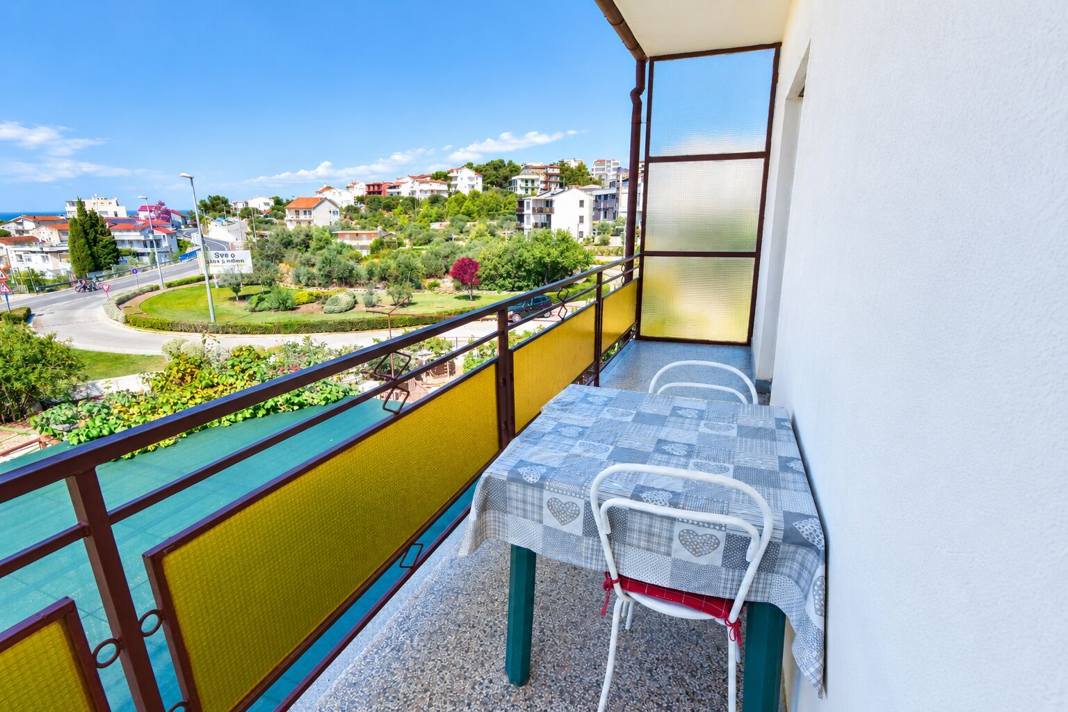 A balcony with a table and chairs, overlooking a residential area with a road and greenery.
