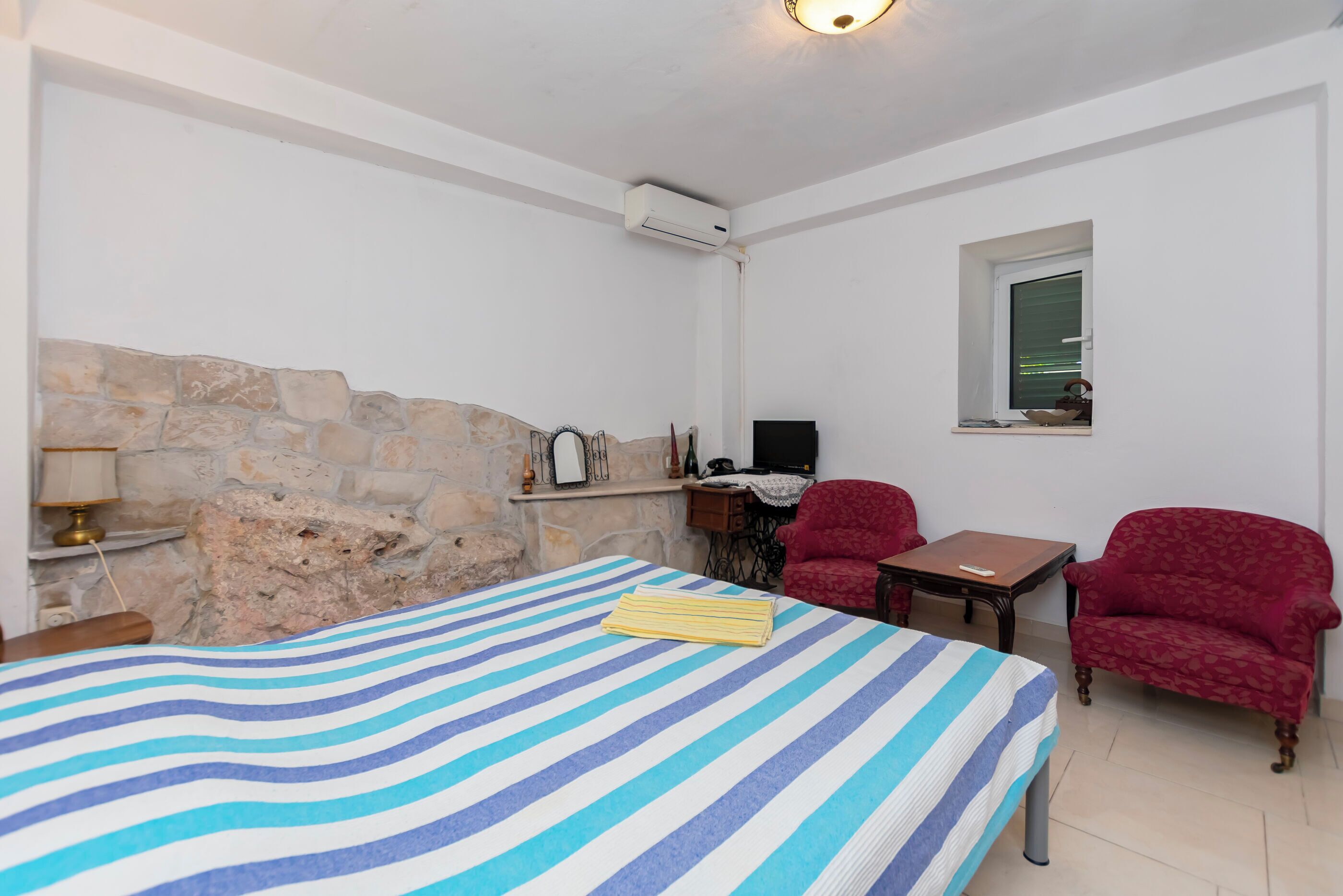 A room with a bed, two red armchairs, a small table, a desk with a computer, a stone wall, and a window.