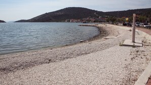 Una spiaggia di ciottoli con un mare tranquillo, una montagna sullo sfondo e una fila di edifici lungo la riva.