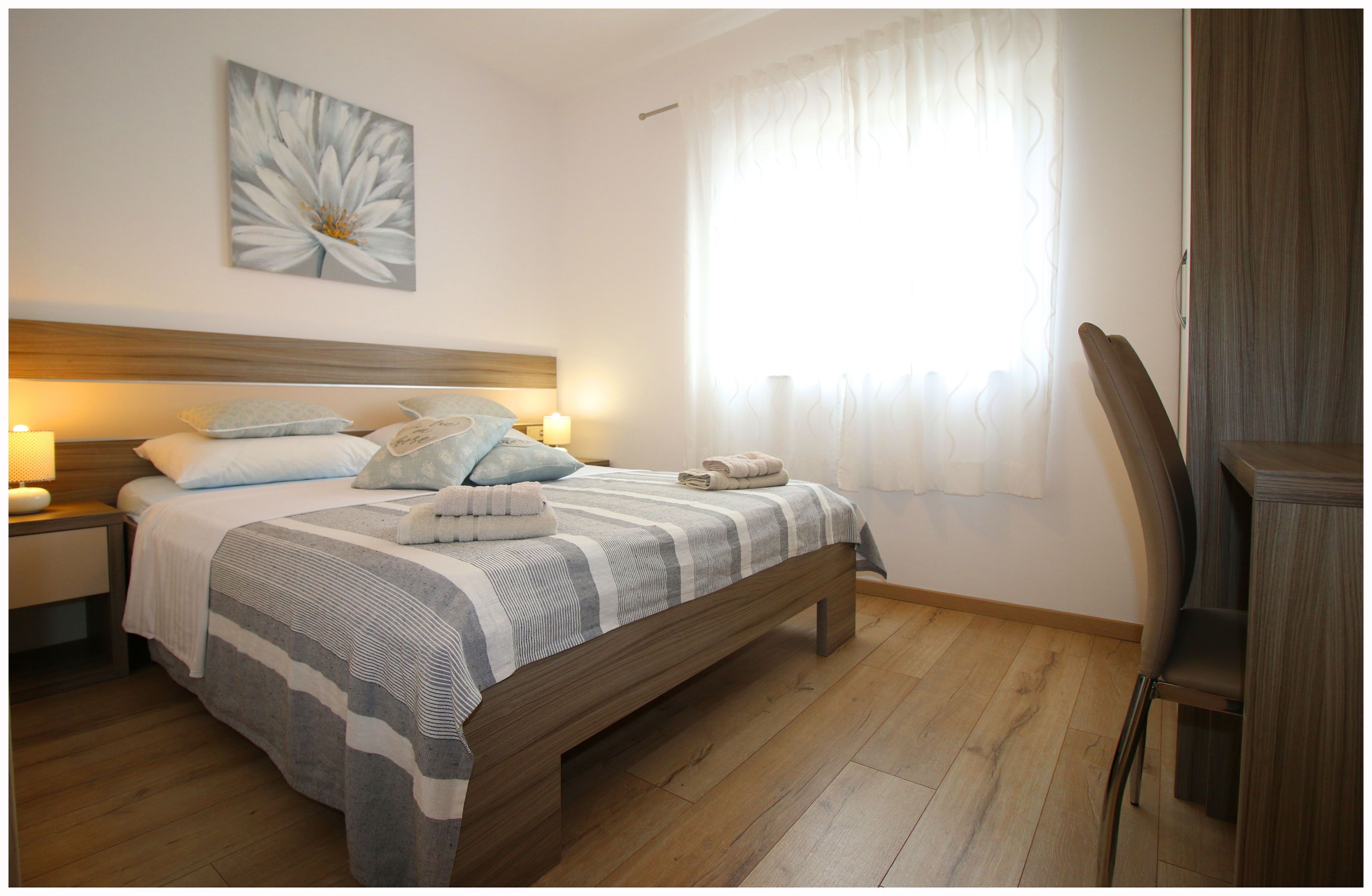A neatly arranged bedroom with a wooden bed, a bedside table with a lamp, a chair, and a large window with curtains.