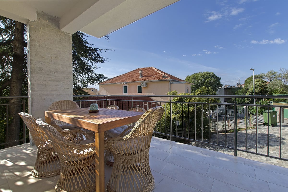 A balcony with a wooden table and wicker chairs, overlooking a building with a red roof.