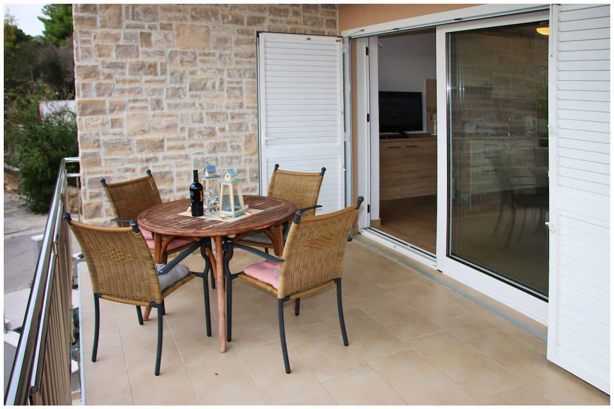 A balcony with a round table and wicker chairs, a glass door leading to an interior space, and a stone wall.