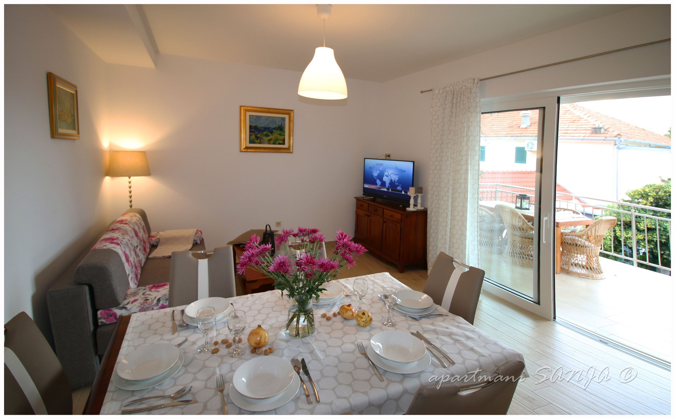A dining area with a table set for four, a sofa, a TV, and a balcony with wicker furniture.