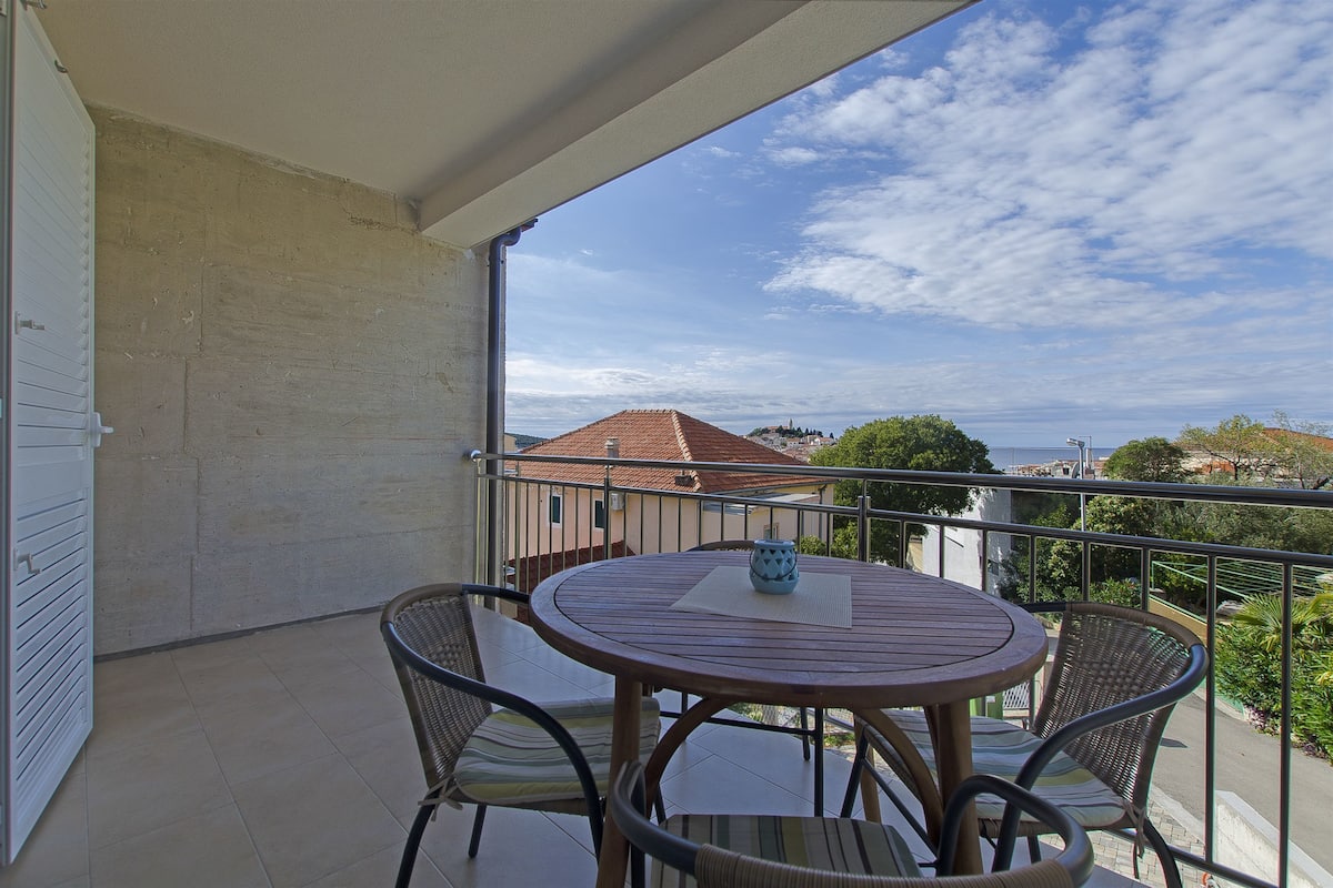 A balcony with a round table and four chairs, overlooking a residential area with a red-roofed building and a view of the sea.