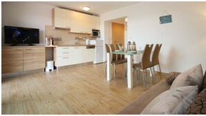 Une cuisine moderne avec des meubles blancs, un four à micro-ondes et une machine à café. On y trouve une table à manger avec des chaises et un plateau en verre. Un canapé, agrémenté de coussins à motifs, se trouve au premier plan.
