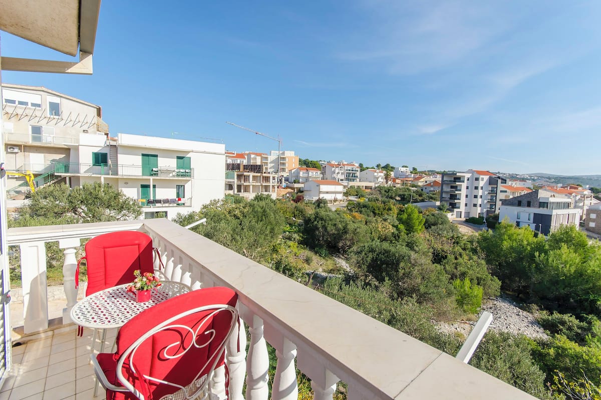 A balcony with red chairs and a table, overlooking a residential area with buildings and greenery.