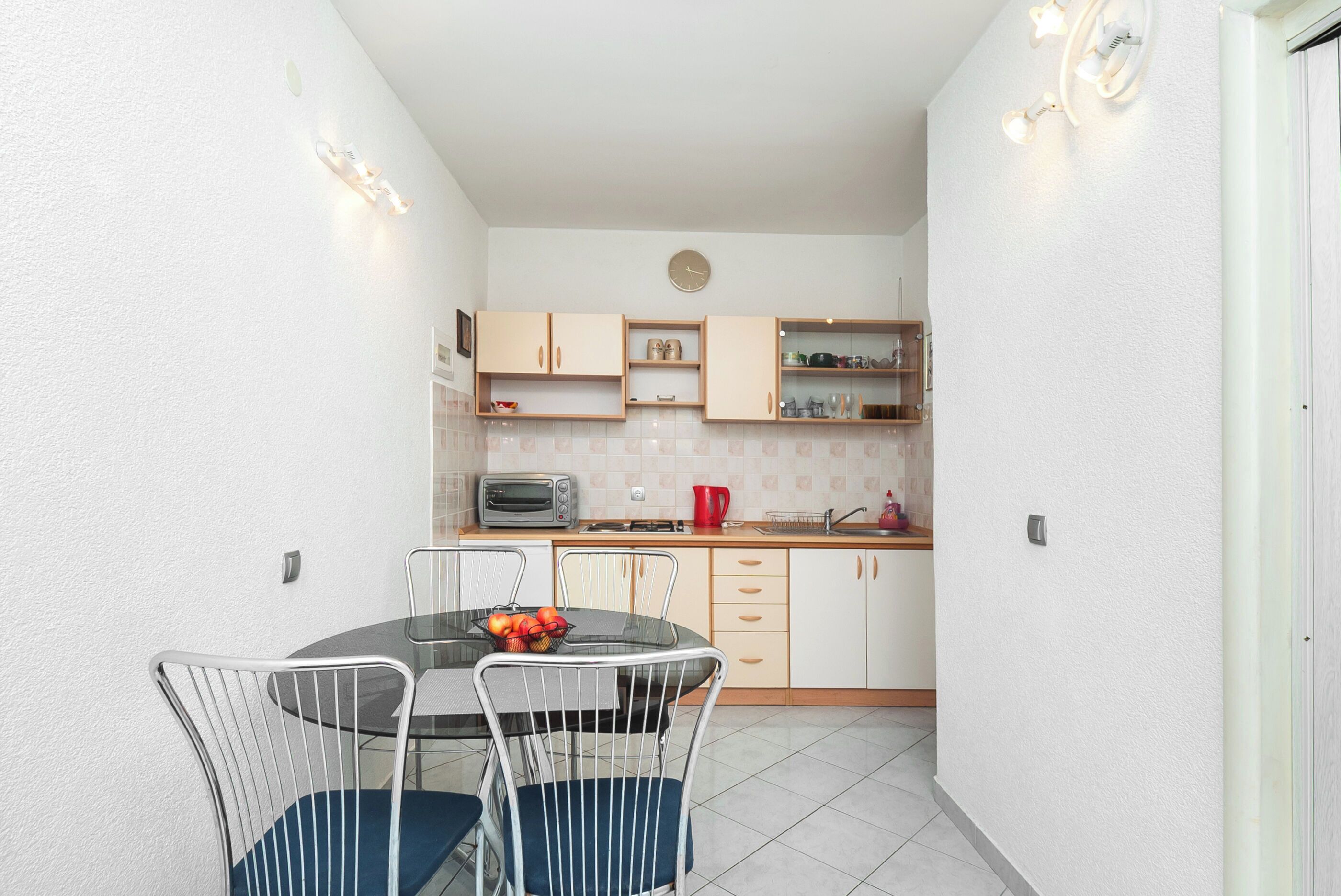 A compact kitchen with a dining area, featuring a round table, chairs, and a fruit bowl.