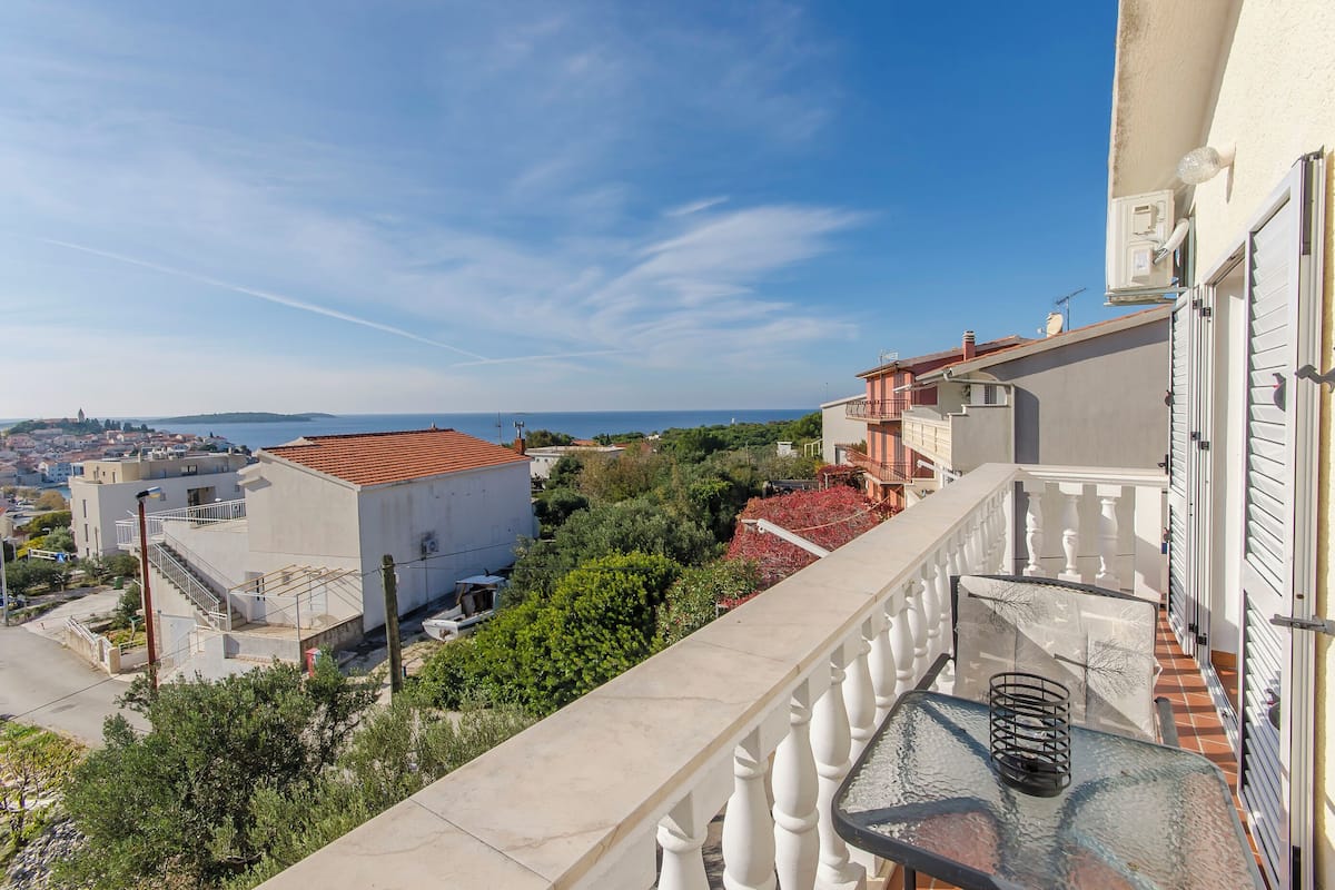 A balcony with a railing, a table with a glass top, and a view of a coastal town with buildings and greenery.