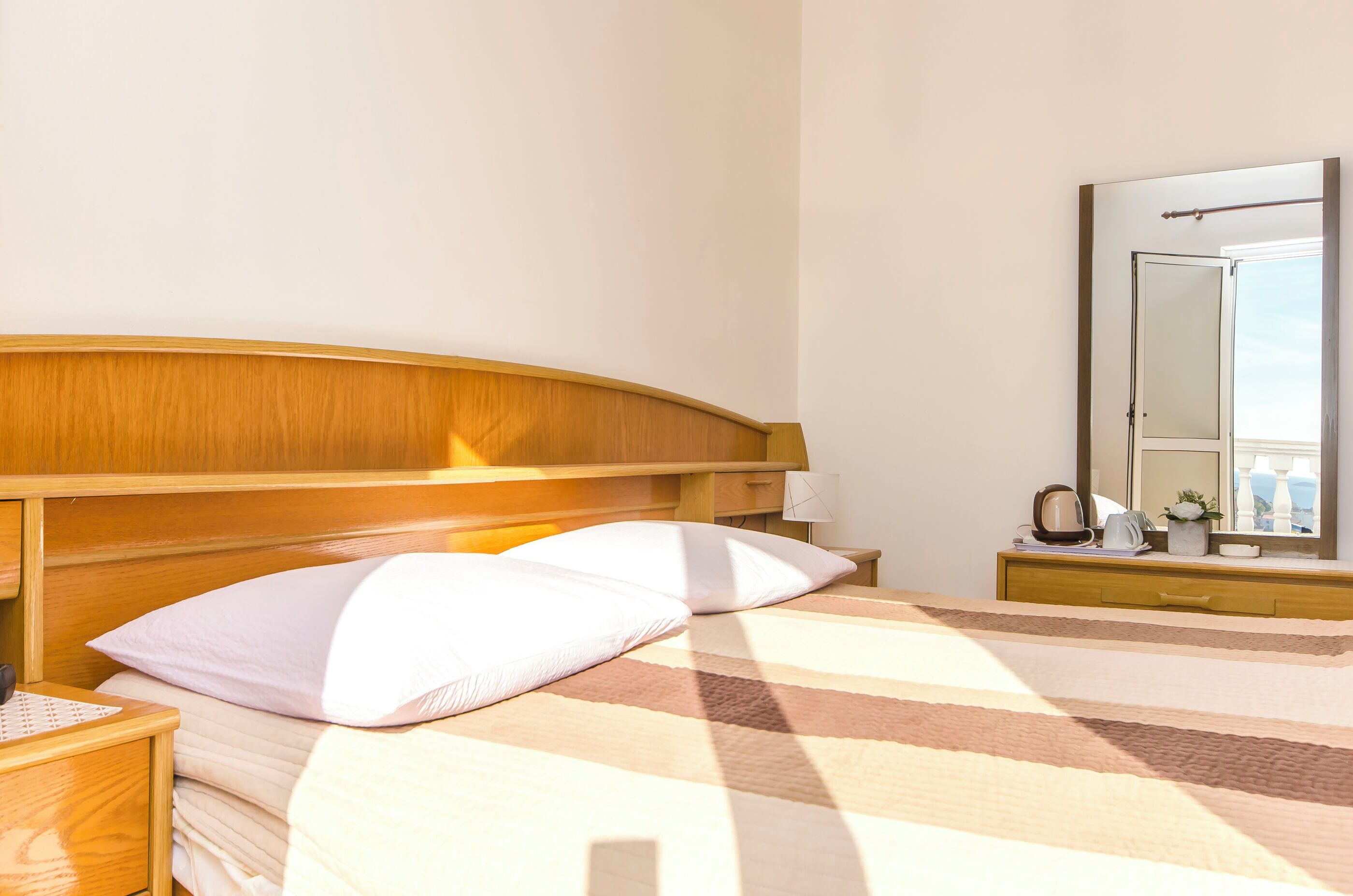 A neatly made bed with a striped blanket, a wooden headboard, a bedside table with a lamp, and a mirror on the wall.