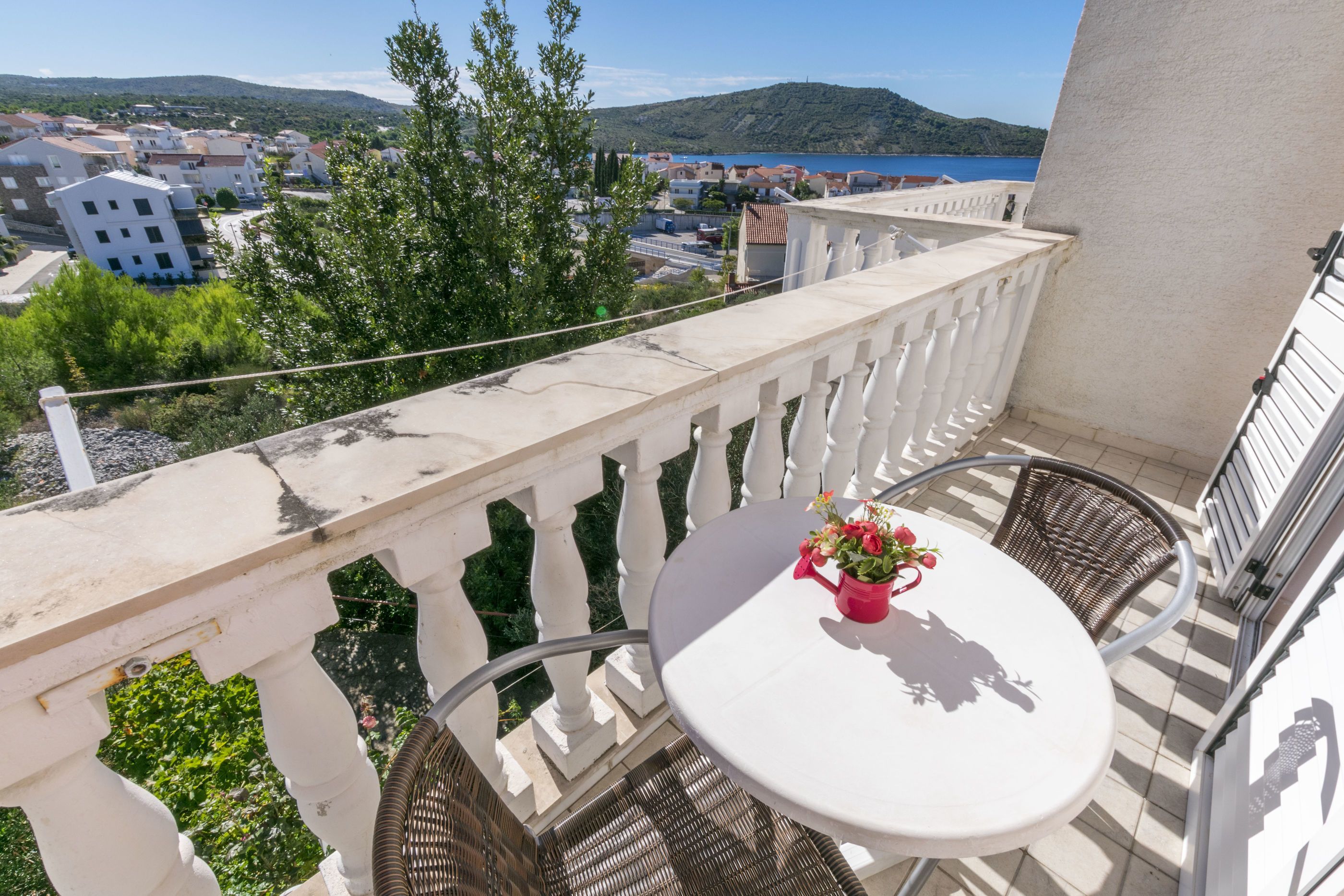 Ein Balkon mit einem weißen Tisch und Korbstühlen, mit Blick auf eine Küstenstadt und das Meer.