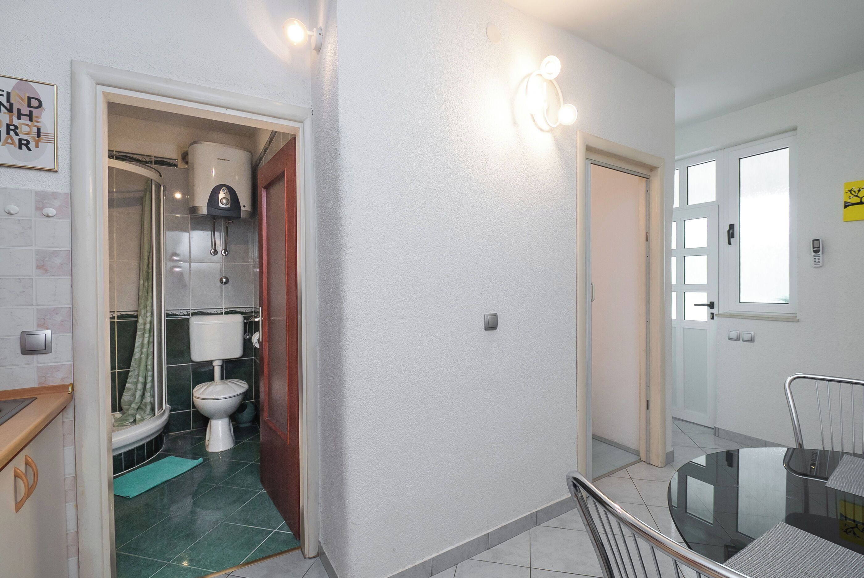 A compact bathroom with a toilet, shower, and a small sink area. There is a dining table with chairs in the adjacent room.
