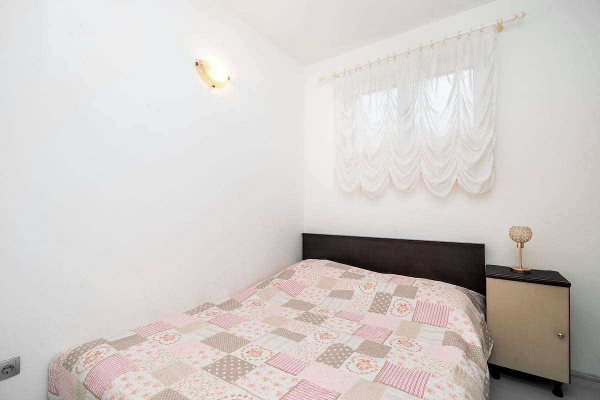 A bedroom with a bed, a nightstand, a wall-mounted light, and a window with a curtain.