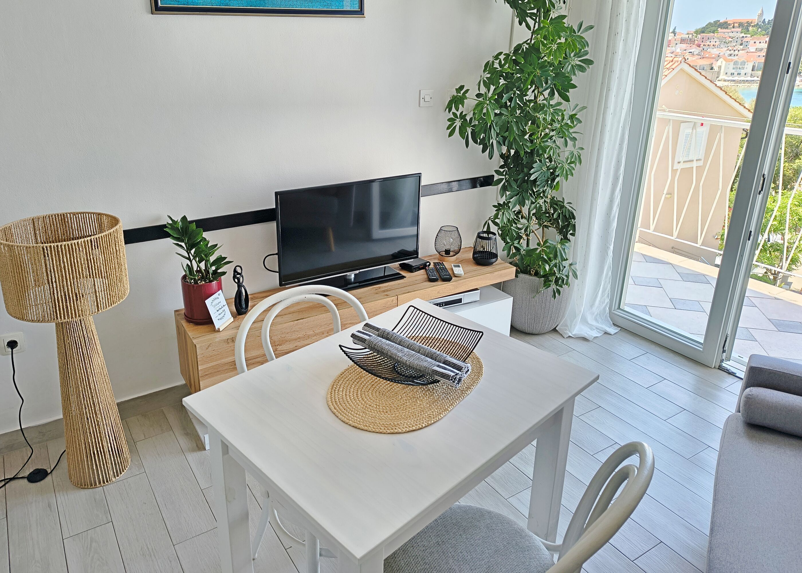 A modern living room with a flat-screen TV, a dining table, chairs, a potted plant, and a balcony with a view.
