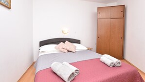A neatly made bed with pink and white bedding, two rolled towels, and a wooden wardrobe in a simple room.