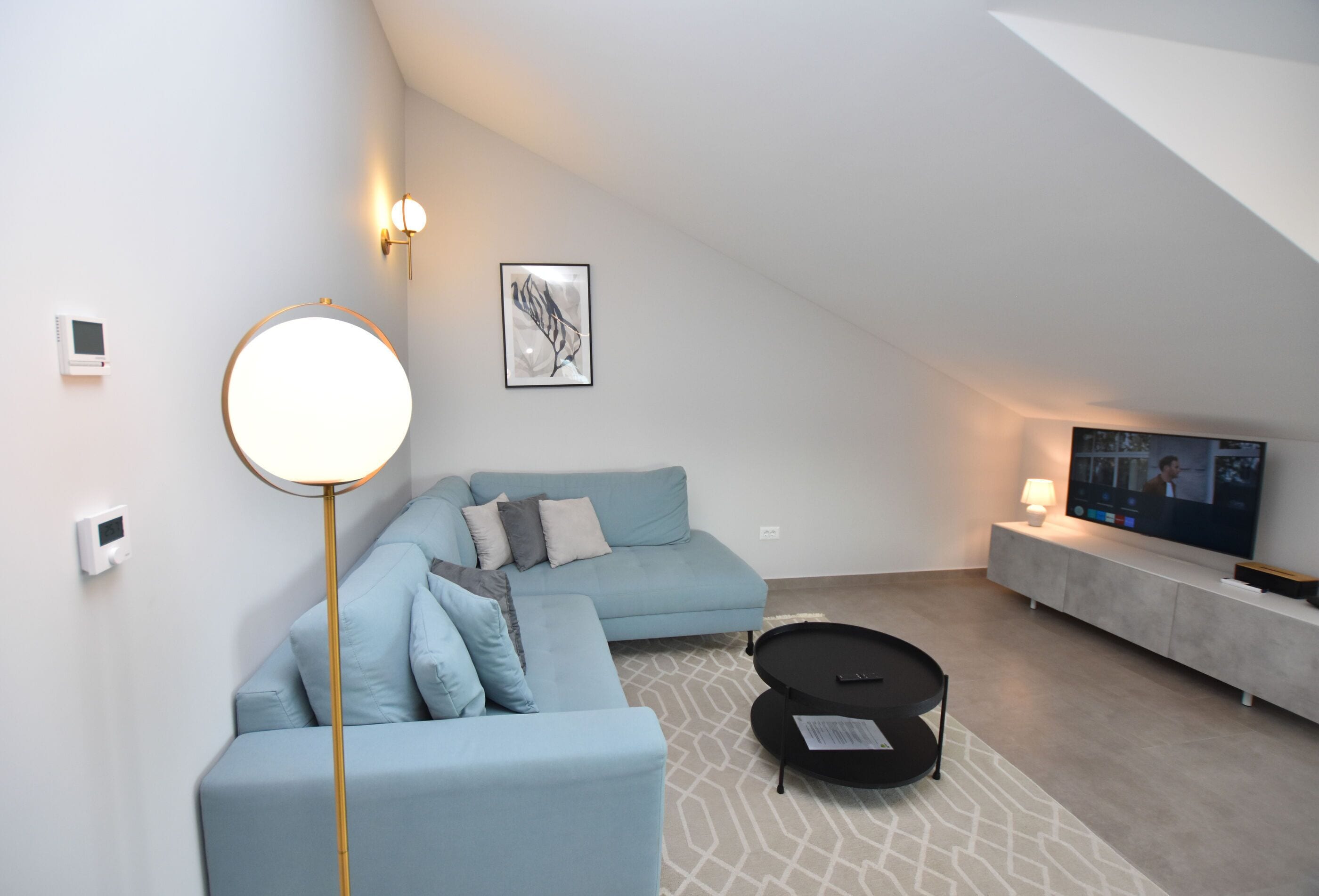 Una sala de estar moderna con un sofá azul claro, una lámpara de pie redonda, una mesa de centro negra y un televisor de pantalla plana sobre un mueble.