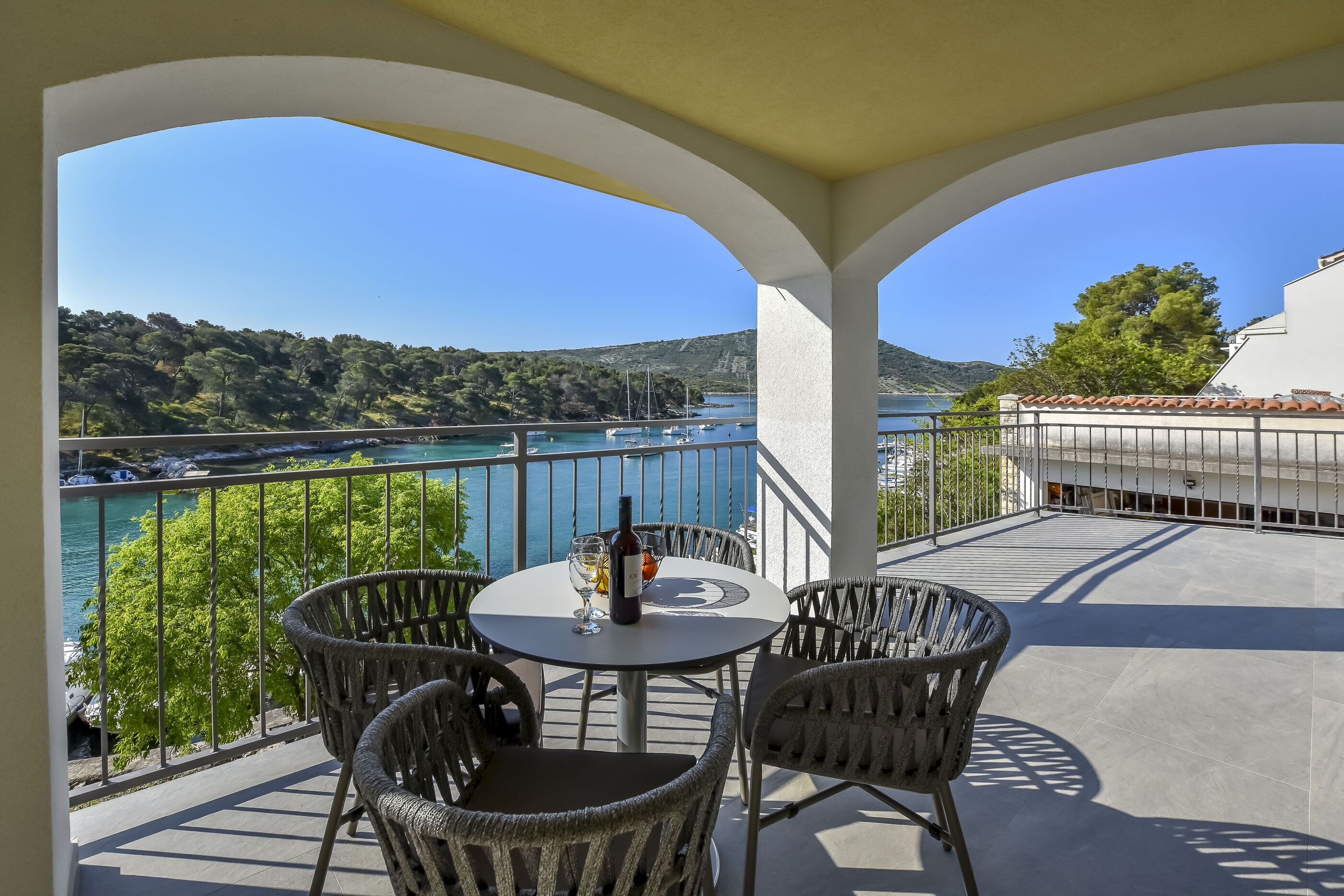 A balcony with wicker chairs and a table, overlooking a scenic coastal view with a clear sky.