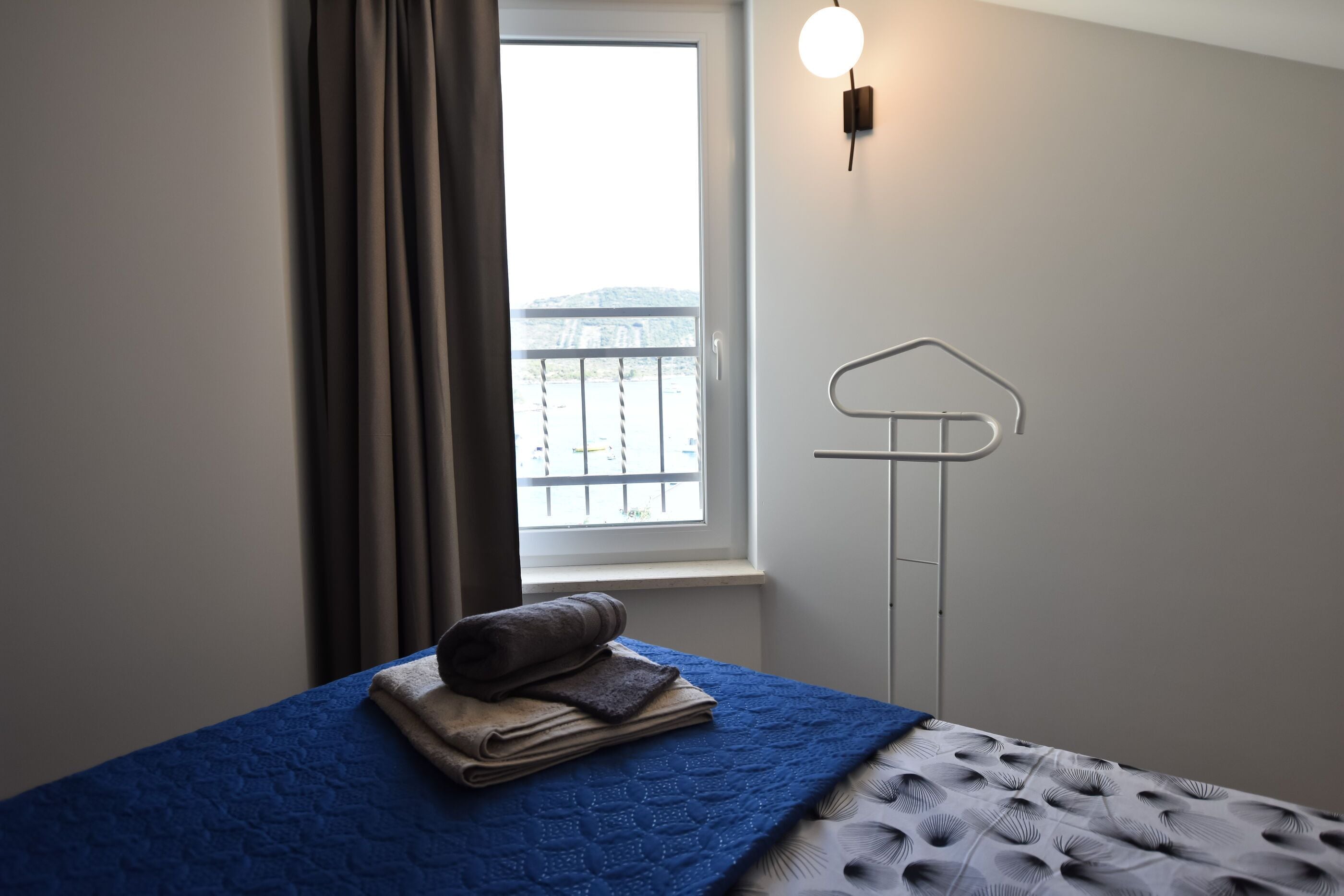 A bedroom with a bed, a window with curtains, a wall-mounted light, and a towel rack.
