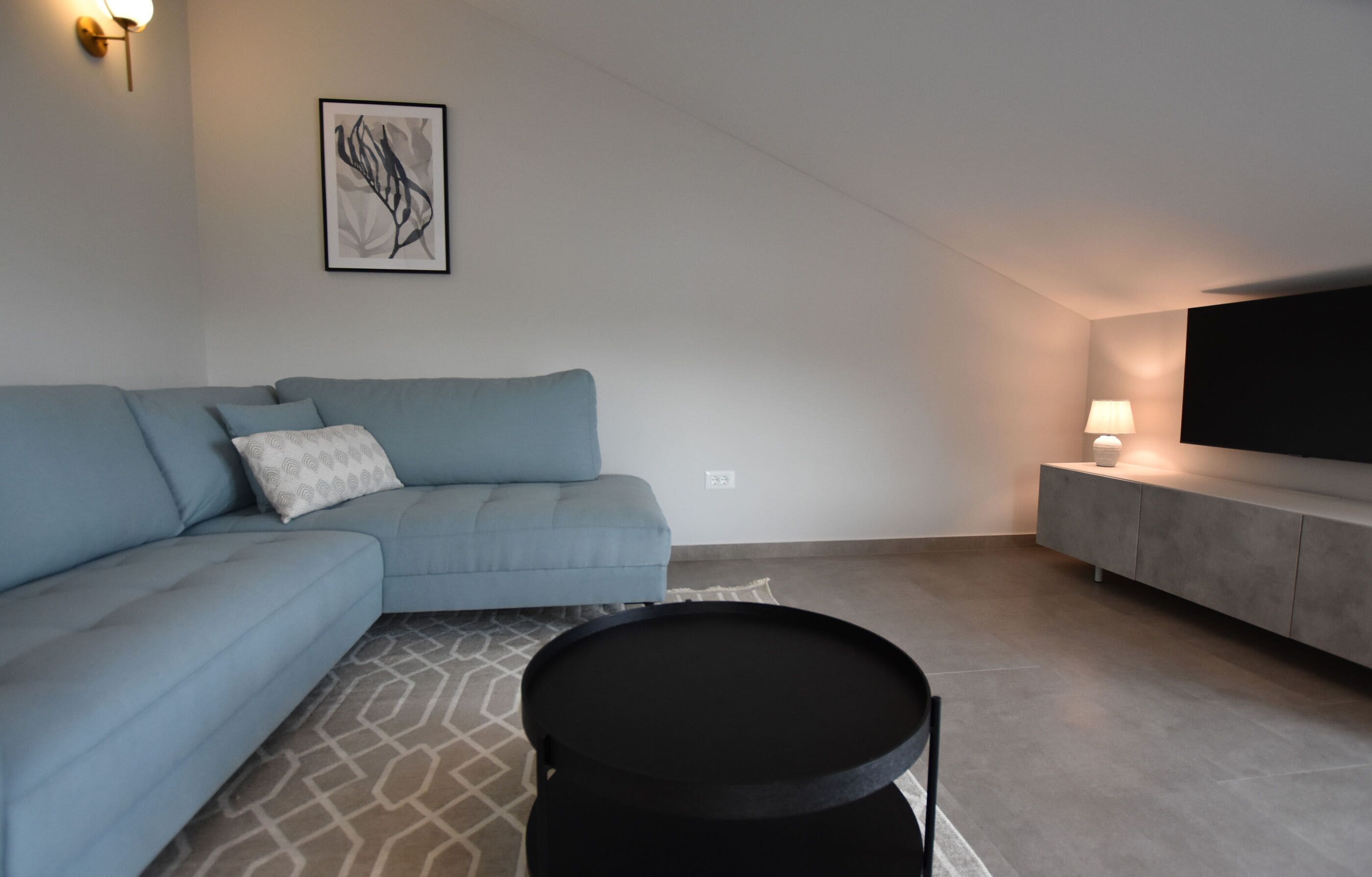 A modern living room with a sectional sofa, a black coffee table, a flat-screen TV on a grey cabinet, and a wall-mounted light.