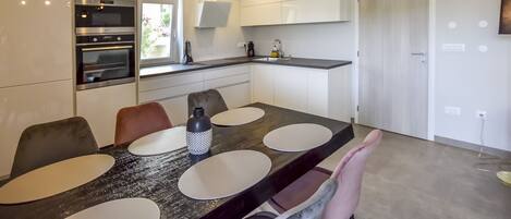 A modern kitchen with a built-in oven, a dining area with a dark wooden table and white placemats, and a pendant light fixture.