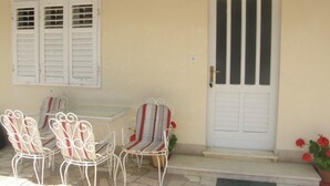 A covered patio with a table and four chairs, a white door with vertical stripes, and a window with shutters. - Apartment Tome (Primosten)