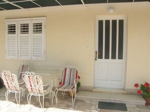 A covered patio with a table and four chairs, a white door with vertical stripes, and a window with shutters. - Apartment Tome (Primosten)