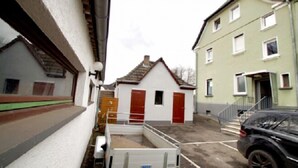 Une ruelle étroite bordée de bâtiments des deux côtés, un petit bâtiment blanc avec des portes rouges, et une voiture noire garée sur le côté droit.